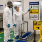 Two food production workers in hairnets and gloves point at hygiene zone and traffic flow signs separating pedestrian and forklift routes inside a processing facility.